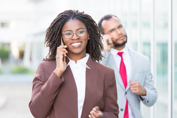 Happy young businesswoman talking on cell outside. Black business woman walking down city street and calling on phone, man speaking on cellphone behind her. Mobile communication concept