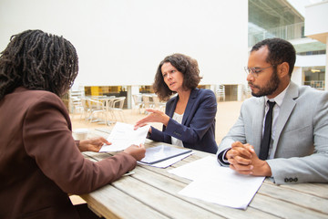 Serious partners discussing agreement terms during meeting in street cafe. Confused business woman pointing at document and asking colleague about contract text. Negotiation concept