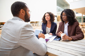 Fototapeta premium Diverse business women discussing deal with partner in cafe. Business man and women sitting at table, reading documents, drinking coffee, using tablet. Deal concept