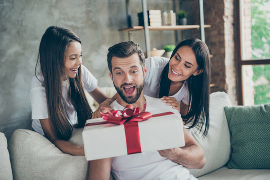 Portrait of nice attractive lovely sweet cheerful cheery excited family wearing casual white t-shirts daddy sitting on sofa mommy giving purchase at industrial style interior living-room indoor