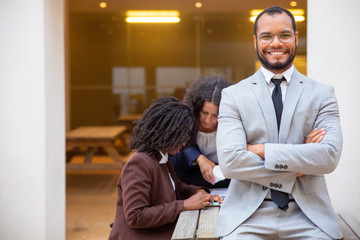Obraz premium Happy positive business man posing with arms crossed in street cafe. His female colleagues sitting and standing at table and talking in background. Successful team concept