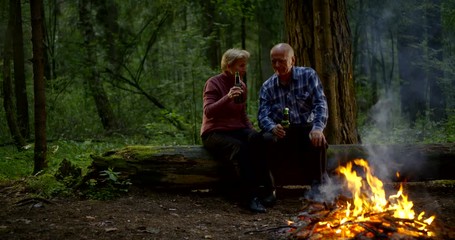 old tourists are sitting in forest and drinking beer from bottles near fire, happy spouses