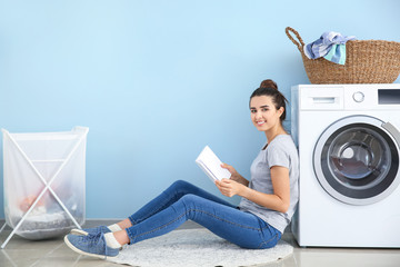 Beautiful young woman reading book while doing laundry at home