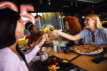 Group of young female friends having fun in restaurant, toasting with wine talking and laughing while dining at table.