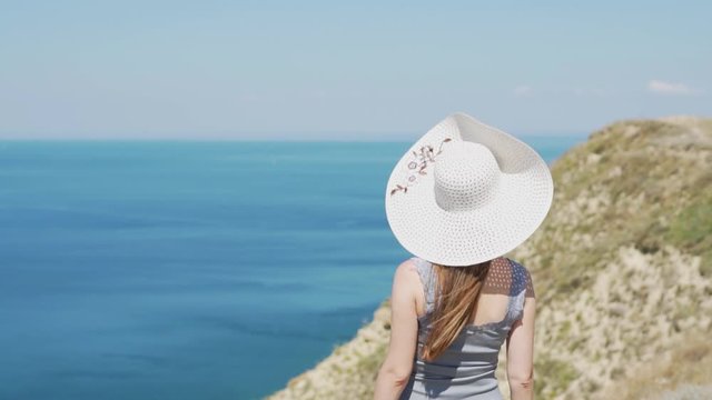 Close - up of European vacation girl in a white hat with a wide brim, rear view. A woman admires a magnificent view of the sea from the height of the mountains in Europe. Rest, relaxation, weekend.