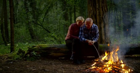 aged woman is hugging her husband in forest, sitting on dry log near campfire