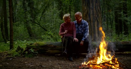 elderly man and woman are looking on fire in forest sitting together and embracing