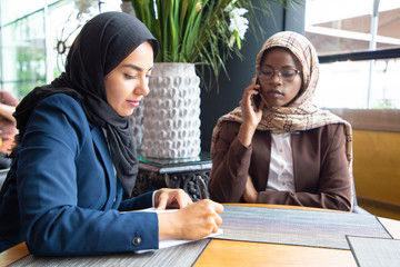 Serious female business colleagues working on project in coffee shop. Muslim business women in...