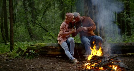 two drunk man and woman are drinking beer sitting near fire in forest