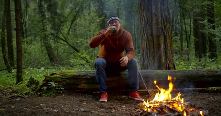 lonely sad man is drinking beer from bottle sitting in forest and looking on flame of campfire