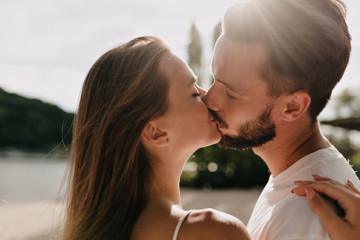 Close up outside portrait of young lovely couple kissing in sunlight on the beach in warm sunny day