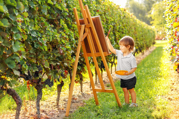 Little talented artist boy painting a picture on easel outdoors at nature in sunny vineyards at bright summer day. Child creativity. Creative hobby and leisure activity. Summer lifestyle.