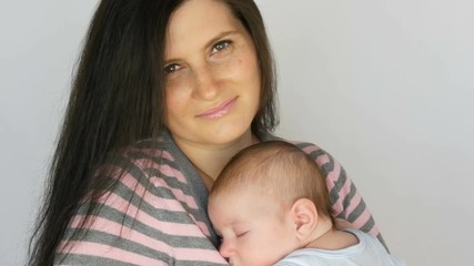 Young beautiful mother with long dark hair is holding a newborn infant baby of two months on a white background in studio