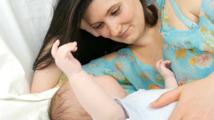 Young beautiful mother with long dark hair is lying on a white bed and playing with a newborn baby of two months