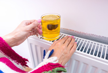 Woman warms herself near a heating radiator and drinks hot tea with lemon in the cold season. The...