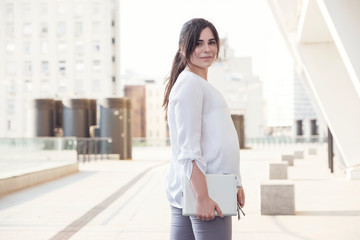 Fototapeta na wymiar Beautiful brunette business woman in white skirt and grey suit trousers working on a tablet in her hands outdoors. European city on background. copy space
