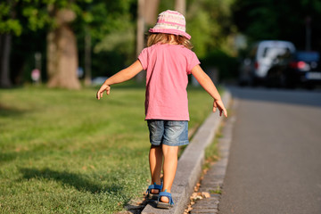 Rear view of a girl child in summer clothing balancing alone on a curb between a lawn and a street...
