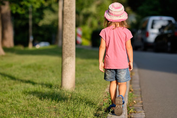 Rear view of a girl child in summer clothing walking alone on a curb between a lawn and a street in the evening light of a summer day
