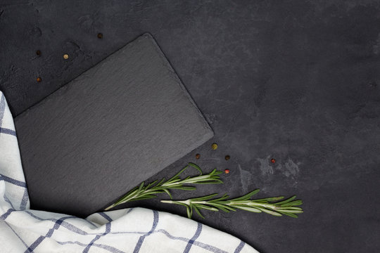 Slate plate, kitchen towel, rosemary, pepper on black concrete background. Cooking, recipe concept. Top view, flat lay, mock up, copy space