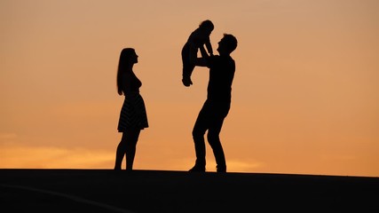 Father rise up little baby and turn around, woman stay near. Young happy family enjoy nice summer day end, shadow figures against yellow sunset clouds on sky, slow motion shot
