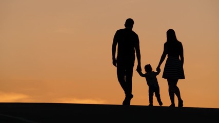 Family with young baby walk against tender sunset sky, full length silhouetted shot. Mother and father holding toddler by hands, little girl make cautious steps. Quiet and peaceful summer evening time