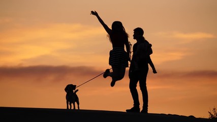 Beautiful portrait, silhouettes of young family against colorful sunset clouds, happy woman jump up high and rise hand. Man hold little baby on hands, dog stay on leash from other side