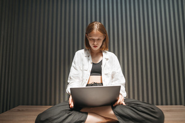Attractive girl in casual casual clothes sits in apartment with laptop on her lap and works against...