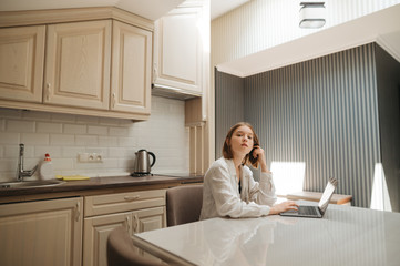 Portrait of a cute girl wearing a white shirt, sitting at a table in the kitchen, using a laptop and looking at the camera. Girl freelancer works at home in apartment on laptop.