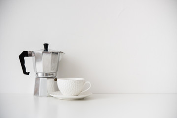 Metal coffee maker and white porcelain cup on table with blank wall