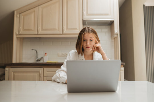Portrait of surprised girl sitting at home in the kitchen at table, looking at laptop screen with shocked face. Surprised girl working on laptop at home in kitchen in apartment, wearing white shirt