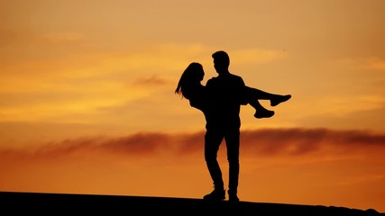 Man holding woman on hands and turn around, silhouetted shot of happy couple against vivid orange sunset sky. Lovely two enjoy life or celebrate some personal event or date