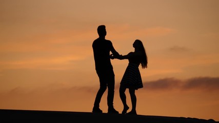 Lovely couple holding hands and turn around, two people dance at sunset. Silhouetted shot against neat yellow sky, nice summer evening time