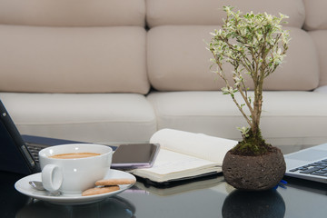 Glass table with a tablet, a cell phones notebook, a cup of coffee and a plant and in the background an elegant cream armchair