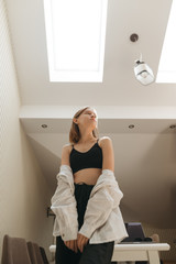 Stylish girl stands in the attic apartment with a bright modern interior and poses for the camera, looking away. Vertical photo.