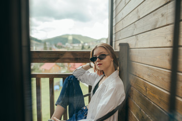 Fototapeta premium Stylish woman in sunglasses sits on a balcony in a country house with a beautiful view,looks in camera with a serious face, wears stylish casual clothes.Girl rests on the balcony in the hotel room
