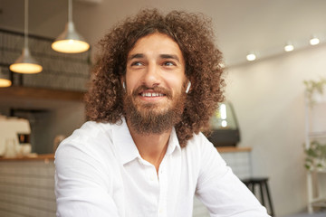 Happy handsome bearded guy with curls looking aside and smiling broadly, being in nice mood, meeting friends during lunch break, posing over coffee house