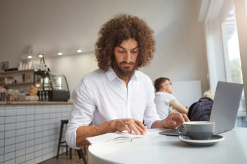 Serious handsome curly guy with beard working remotely with his laptop at coffee shop, looking intently in his notes and wrinkling forehead