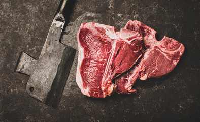 Flat-lay of fresh uncooked beef meat t-bone steaks and butchers cleaver over rough black stone...