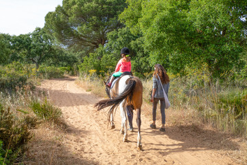 view from behind of four years old blonde girl, with equestrian cap, riding a horse next to her mother walking on track in a forest