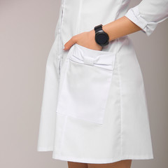 close-up young stylish nurse with one hand with wrist watch in the pocket of her medical dress on white wall background. medical fashion concept. free space