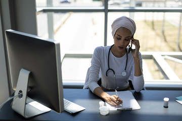 Beautiful mixed-race female doctor talking on mobile phone while writing on clipboard