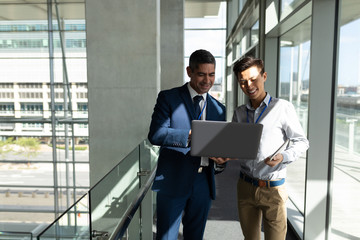 Fototapeta premium Front view of two businessman standing on the first floor walkway and working on a laptop in office