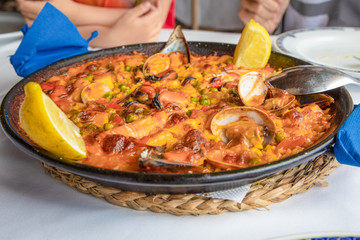 close up view of typical Spanish paella with seafood (mussel, prawn, lobster, squid, clam), rice and lemon ready to serve on white tablecloth of restaurant