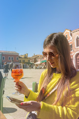 Young woman in yellow jacker with glass of aperol sitting on square in sunny day. Burano, Venice, Italy.