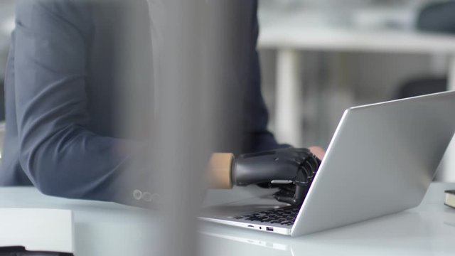 Close up shot of unrecognizable businessman with prosthetic bionic hand typing on laptop at office desk