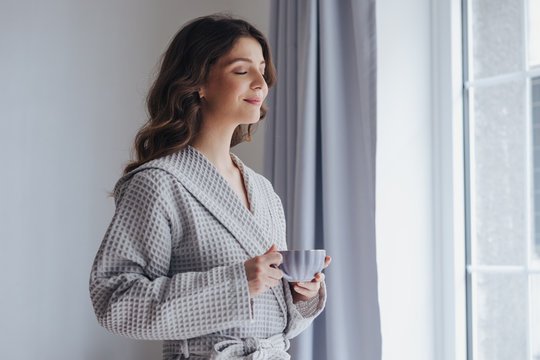 Portrait of pretty woman in bathrobe enjoying morning standing by the window and holding cup of tea.