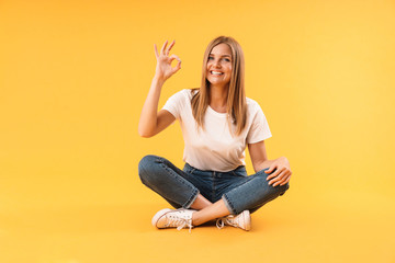 Image of charming blond woman smiling and showing ok sign while sitting with legs crossed