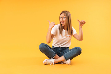 Image of young blond woman rejoicing and pointing fingers at copyspace while sitting with legs crossed