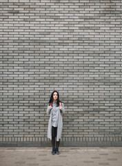 Beautiful attractive young woman with a backpack standing half a turn near the gray brick wall. Lonely look. Pensive girl. Copy space.