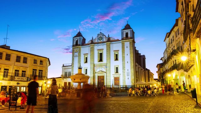 Evora, Portugal. View of Roman Catholic Cathedral of Evora, Portugal at sunset. Illuminated historical buildings at the square with motion blurred people. Time-lapse of sky gettind dark, zoom in
